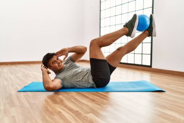 Genç İspanyol adam gülümsüyor. Spor merkezinde top kullanarak karın kasları çalıştırıyor.