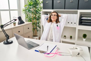 Young woman wearing doctor uniform relaxed with hands on head at clinic