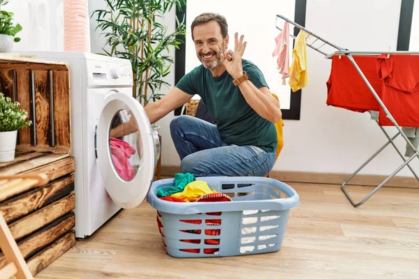 髭を生やした中年の男が洗濯機に洗濯機を入れて指でOkサインをし 優しいジェスチャーを笑顔優れたシンボル — ストック写真