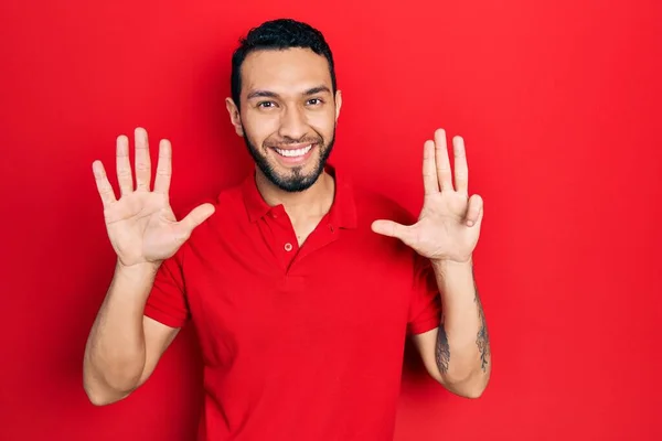 自信と幸せ笑みを浮かべている間 髭を生やしたヒスパニック系の男は カジュアルな赤いTシャツを着て表示され 指の番号9で指摘 — ストック写真