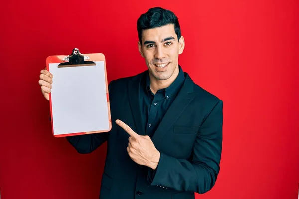 手と指で幸せなポインティングとクリップボードを保持しているハンサムなヒスパニック系ビジネス男 — ストック写真