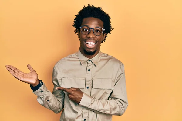 手で提示し 指で指している間 カジュアルな服や眼鏡を身に着けている若いアフリカ系アメリカ人の男性は カメラに驚いて笑顔 — ストック写真
