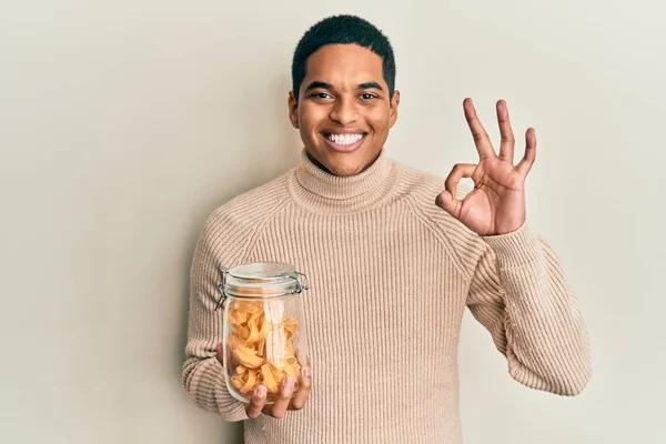 Genç, yakışıklı İspanyol adam elinde makarna kavanozuyla tamam işareti yapıyor, gülümseyerek mükemmel bir sembol oluşturuyor. 