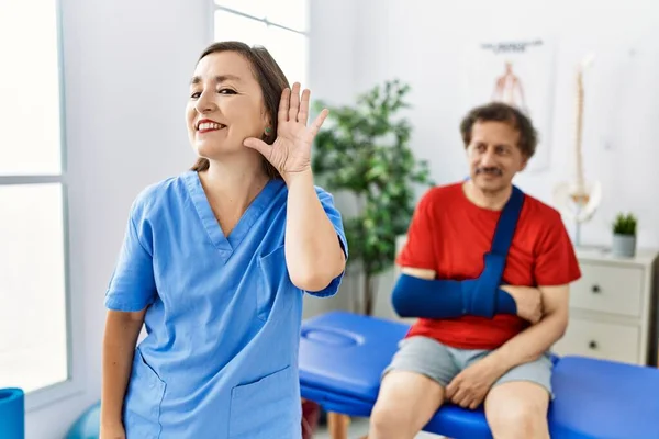 Middle age doctor woman with patient with arm injury at rehabilitation clinic smiling with hand over ear listening and hearing to rumor or gossip. deafness concept.