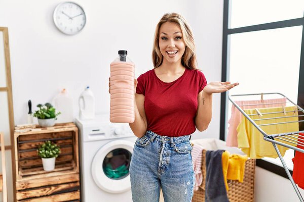 Young caucasian woman holding detergent bottle at laundry room celebrating achievement with happy smile and winner expression with raised hand 