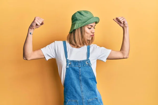 年轻的高加索金发女人穿着斜纹棉布连衣裙 头戴90年代风格的帽子 露出手臂肌肉的笑容 感到自豪 健身概念 — 图库照片