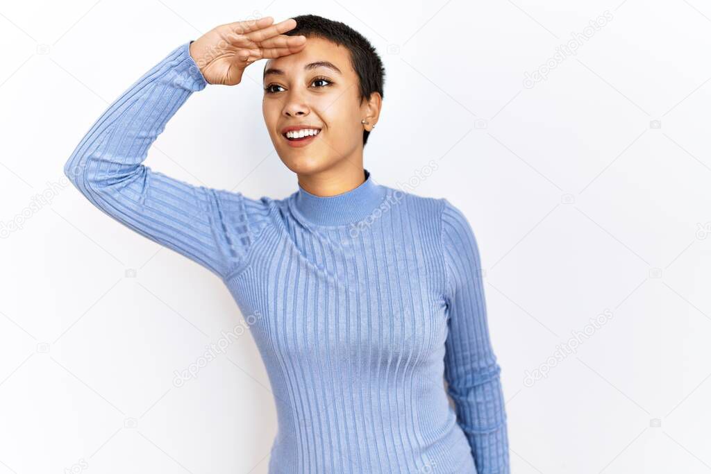 Young hispanic woman with short hair standing over isolated background very happy and smiling looking far away with hand over head. searching concept. 