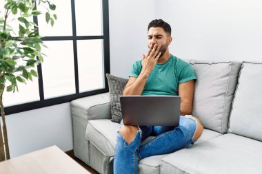 Young handsome man with beard using computer laptop sitting on the sofa at home bored yawning tired covering mouth with hand. restless and sleepiness. 