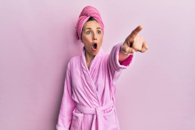 Young blonde woman wearing shower towel cap and bathrobe pointing with finger surprised ahead, open mouth amazed expression, something on the front 