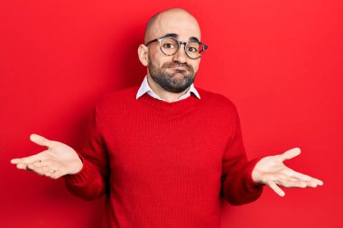 Sıradan kıyafetler giyen, gözlüklü, kafası karışmış, elleri ve kolları havada genç kel bir adam. şüphe kavramı. 