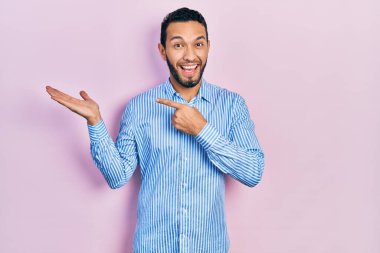 Hispanic man with beard wearing casual blue shirt amazed and smiling to the camera while presenting with hand and pointing with finger. 