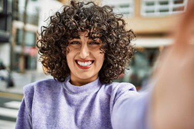 Orta Doğulu genç bir kadın gülümsüyor. Sokakta kameranın önünde özçekim yapıyor.