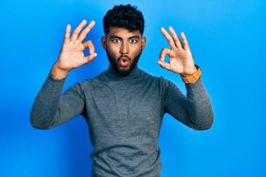 Arab man with beard wearing turtleneck sweater looking surprised and shocked doing ok approval symbol with fingers. crazy expression 