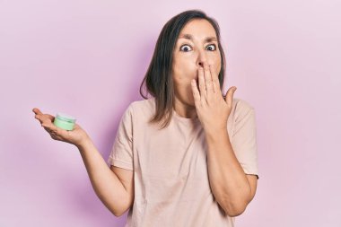 Orta yaşlı İspanyol kadın elinde kozmetik nemlendirici krem tutarak ağzını eliyle kapatıyor, şok geçiriyor ve hata yapmaktan korkuyor. şaşırmış ifade 