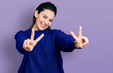 Gündelik kıyafetler giymiş, dili açık gülümseyen, zafer işareti yapan genç İspanyol kadın. İki numara.. 