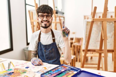 Hispanic man with beard at art studio showing and pointing up with finger number one while smiling confident and happy. 