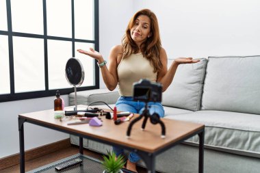 Orta yaşlı İspanyol kadın evinde akıllı telefonuyla makyaj yapıyor ve elleri ve kolları havada, kafası karışmış bir ifade takınıyor. şüphe kavramı. 