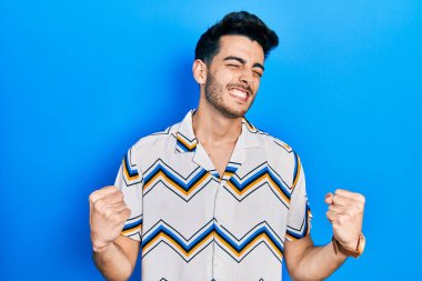 Gündelik kıyafetler giyen genç İspanyol adam çok mutlu ve heyecanlı. Kazanan jestler yapıyor. Kolları havada, gülümsüyor ve başarı için bağırıyor. kutlama konsepti. 
