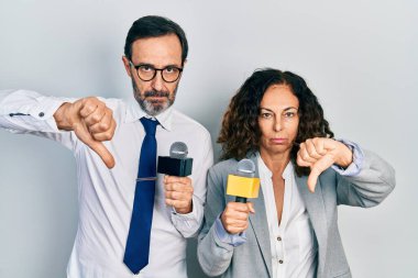 Middle age couple of hispanic woman and man holding reporter microphones with angry face, negative sign showing dislike with thumbs down, rejection concept 