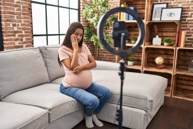 Young pregnant woman recording vlog tutorial with smartphone at home thinking looking tired and bored with depression problems with crossed arms. 