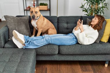 Beyaz bir kadın akıllı telefon kullanıyor. Kanepede köpeğiyle yatıyor.