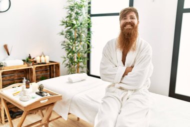 Young redhead man wearing bathrobe sitting on massage table at beauty center