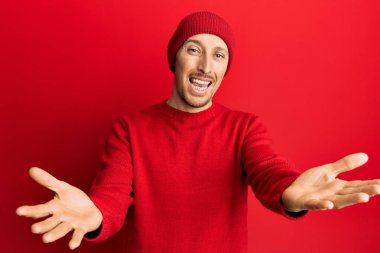 Bald man with beard wearing wool sweater and winter hat looking at the camera smiling with open arms for hug. cheerful expression embracing happiness. 