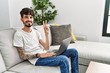 Hispanic man with beard sitting on the sofa showing and pointing up with fingers number two while smiling confident and happy. 