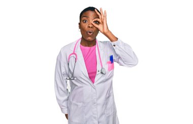 Young african american woman wearing doctor uniform and stethoscope doing ok gesture shocked with surprised face, eye looking through fingers. unbelieving expression. 