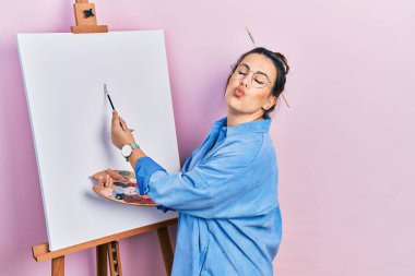 Young hispanic woman standing drawing with palette by painter easel stand looking at the camera blowing a kiss being lovely and sexy. love expression. 