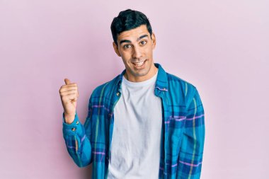 Yakışıklı İspanyol bir adam, günlük kıyafetler giyiyor, gülümsüyor, gülümsüyor ve başparmağıyla yan tarafı işaret ediyor.. 