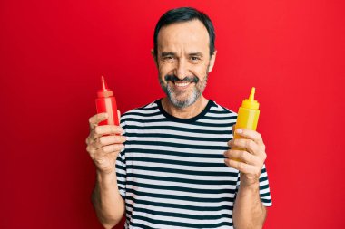 Middle age hispanic man holding ketchup and mustard bottle smiling with a happy and cool smile on face. showing teeth. 