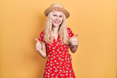 Sarı saçlı, yaz şapkası takan güzel beyaz bir kadın el hareketleriyle olumlu bir hareket yapıyor, gülümsüyor ve mutlu. Neşeli ifade ve kazanan jest. 