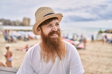 Genç, kızıl saçlı, gülümseyen turist kumsaldaki bankta mutlu bir şekilde oturuyor..