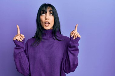 Young brunette woman with bangs wearing turtleneck sweater amazed and surprised looking up and pointing with fingers and raised arms. 