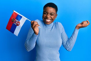 Young african american woman holding serbia flag screaming proud, celebrating victory and success very excited with raised arm 