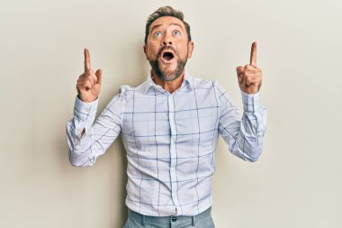 İş kıyafetleri giyen orta yaşlı bir adam şaşırmış ve şaşırmış bir şekilde yukarı bakıyor ve parmaklarıyla işaret ediyor ve kolunu kaldırıyor.. 