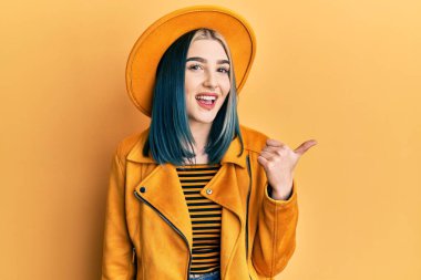 Sarı şapkalı, deri ceketli, mutlu yüzlü gülümseyen ve baş parmağıyla yan tarafı gösteren modern bir kız.. 