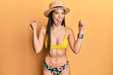 Young hispanic woman wearing bikini and summer hat celebrating surprised and amazed for success with arms raised and open eyes. winner concept. 