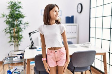 Young hispanic girl standing at pediatrician clinic looking away to side with smile on face, natural expression. laughing confident. 