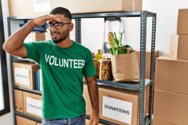 Young indian man volunteer holding donations box very happy and smiling looking far away with hand over head. searching concept. 