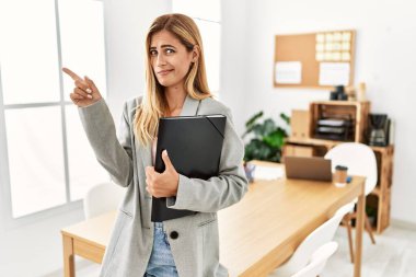 Blonde business woman at the office pointing aside worried and nervous with forefinger, concerned and surprised expression 