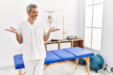 Orta yaşlı İspanyol terapist. Ağrı kesici kliniğinde çalışıyor. Gülümsüyor, iki elini de açık tutuyor. 