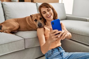 Kafkasyalı genç bir kadın akıllı telefonun yanında evde köpekle oturmuş selfie çekiyor.