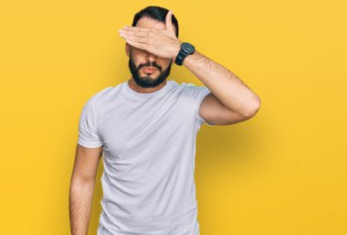 Sakallı genç adam, sıradan beyaz tişörtler giyiyor, gözleri elle örtüyor, ciddi ve üzgün görünüyor. Görme, saklama ve reddetme konsepti 