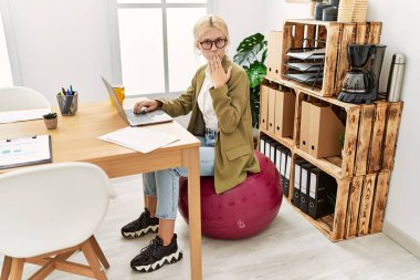 Ofiste çalışan beyaz, güzel bir iş kadını pilates topunun üzerine oturup ağzını eliyle kapatıyor, şok geçiriyor ve hata yapmaktan korkuyor. şaşırmış ifade 