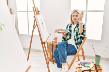 Young artist woman painting on canvas at art studio doing italian gesture with hand and fingers confident expression 
