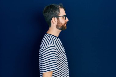 Sakallı, çizgili t-shirt ve gözlüklü, rahat bir profil pozu veren, kendine güveni olan beyaz bir adam.. 