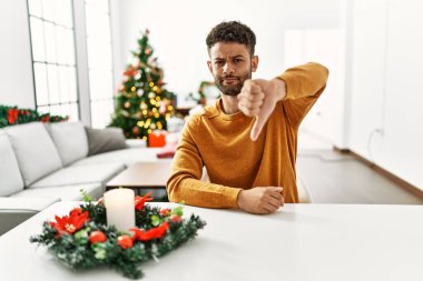 Arap genç adam Noel ağacının yanında masada oturuyor. Mutsuz ve kızgın görünüyor. Reddedilmiş ve olumsuz bir hareket sergiliyor. kötü ifade. 