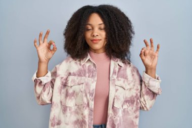 Genç Afrikalı Amerikalı kadın mavi arka planda duruyor rahatla ve gülümse gözleri kapalı parmaklarıyla meditasyon hareketi yapıyor. yoga konsepti. 